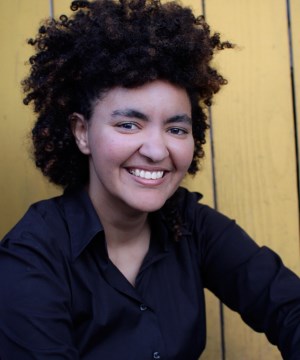 Image description: A sitting and grinning soft-butch woman with a gigantic afro and sides pulled back wears business black shirt, unbuttoned on top, and leans her left arm on her knee outside of the picture and her blue jean barely visible, in front of a mustard yellow background.